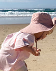 A toddler wearing a light pink sun hat and matching pink shirt stands barefoot on a sandy beach, looking down toward the sand with the ocean and blue sky in the background.