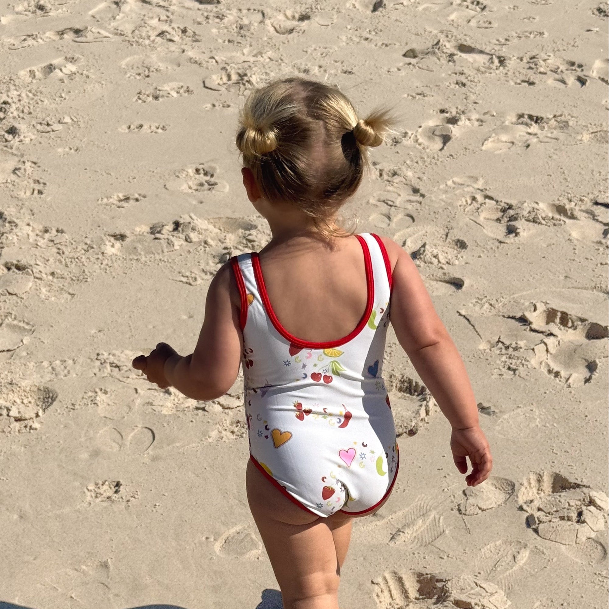 A toddler walking on a sandy beach from behind, wearing a white one-piece swimsuit with red trim and a small multicolor fish pattern.
