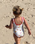 A toddler walking on a sandy beach from behind, wearing a white one-piece swimsuit with red trim and a small multicolor fish pattern.