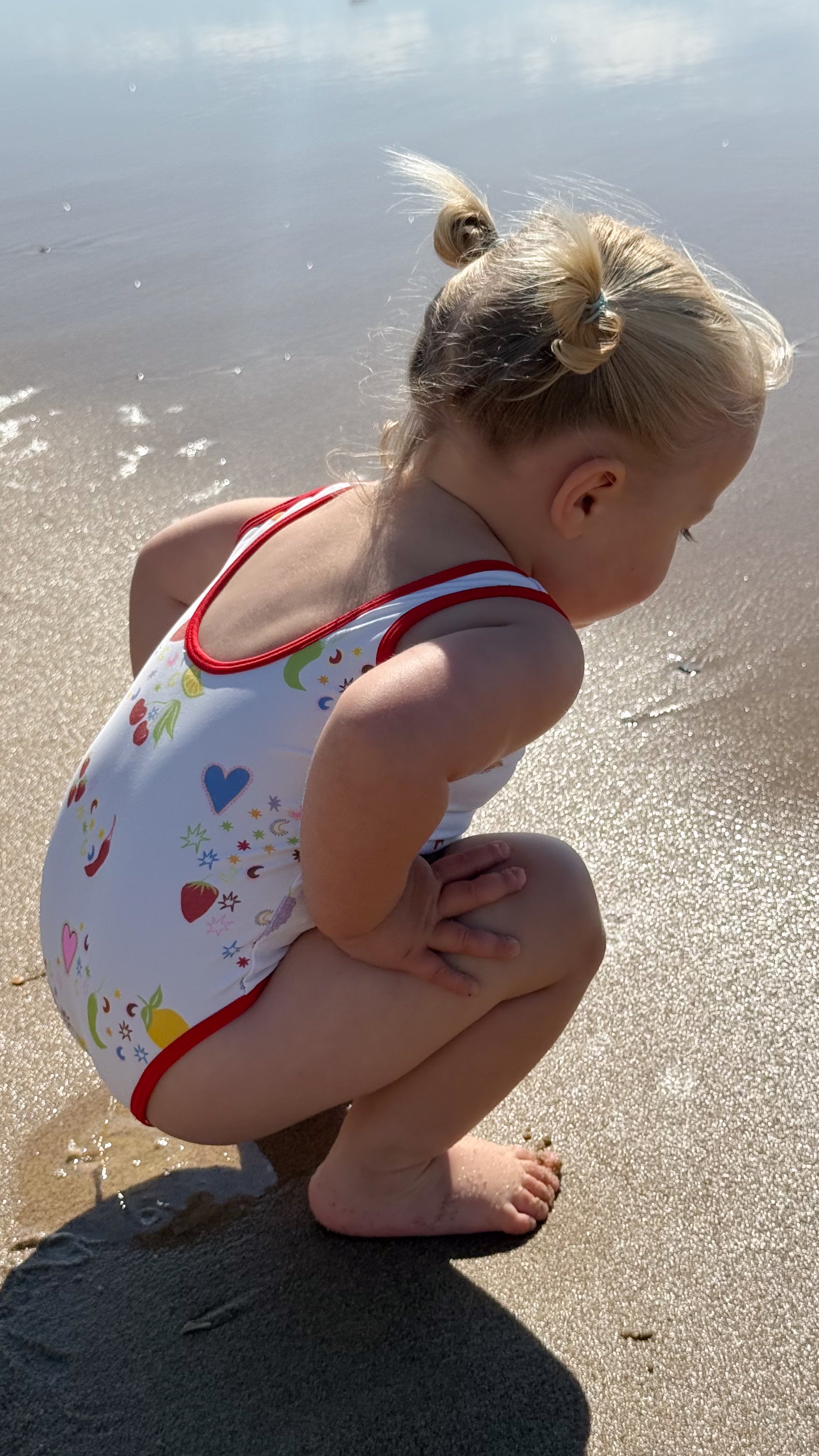 A toddler girl crouching on a sandy beach wearing a white one-piece swimsuit with red trim and a colorful heart and strawberry pattern.
