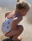 A toddler girl crouching on a sandy beach wearing a white one-piece swimsuit with red trim and a colorful heart and strawberry pattern.