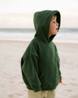 A young child standing on a sandy beach wearing a dark green hooded sweatshirt and light khaki pants.