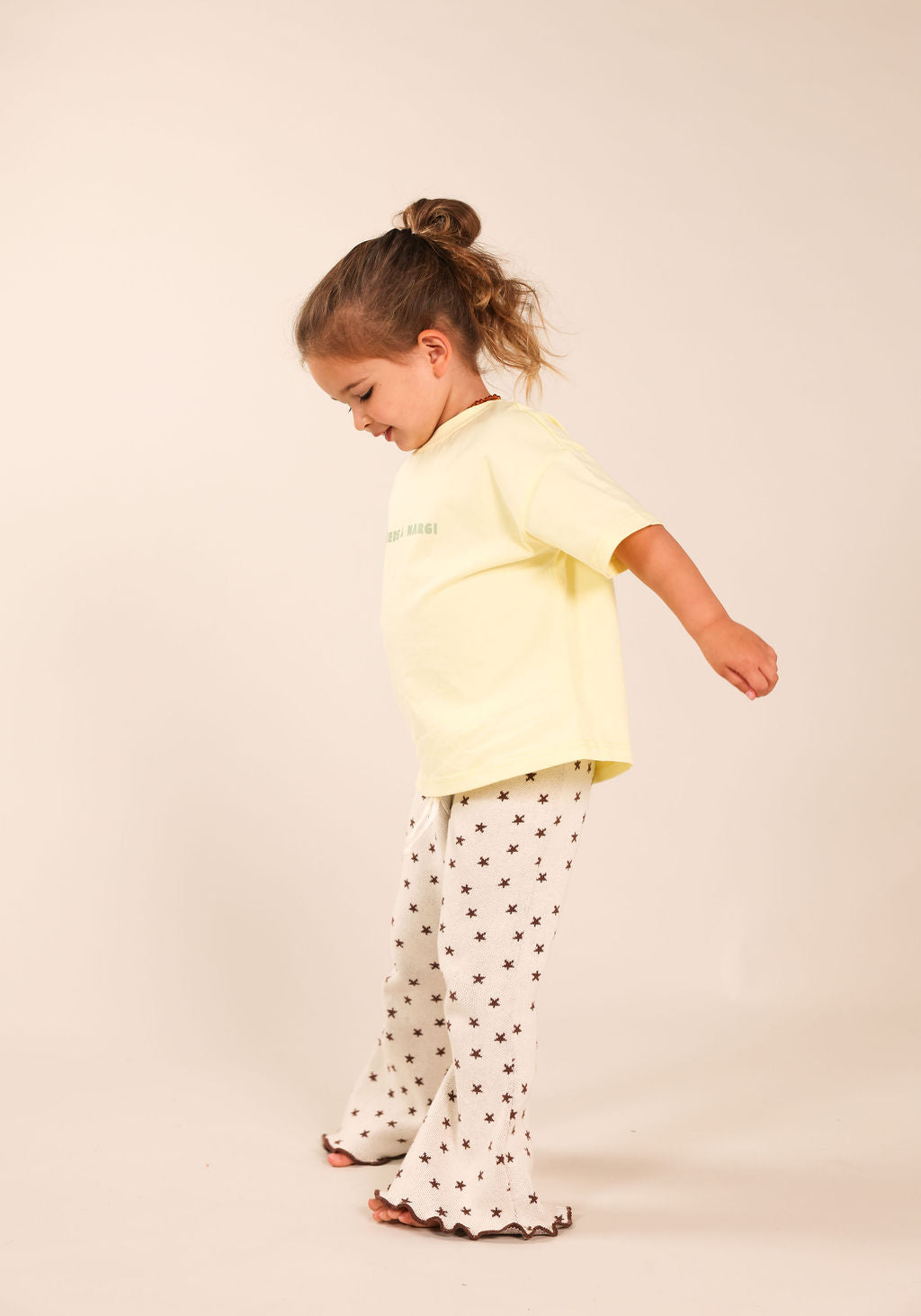 A young child wearing a pale yellow short-sleeve t-shirt and white flared pants with small dark star prints, standing against a plain beige studio background.