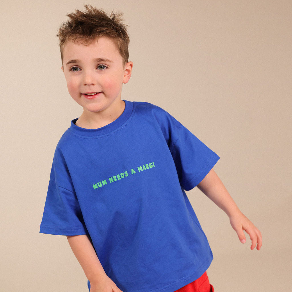A young boy modeling a bright blue crewneck t-shirt with green text that reads 'HUN NEEDS A MARGI!' and red pants, standing against a solid beige studio background.