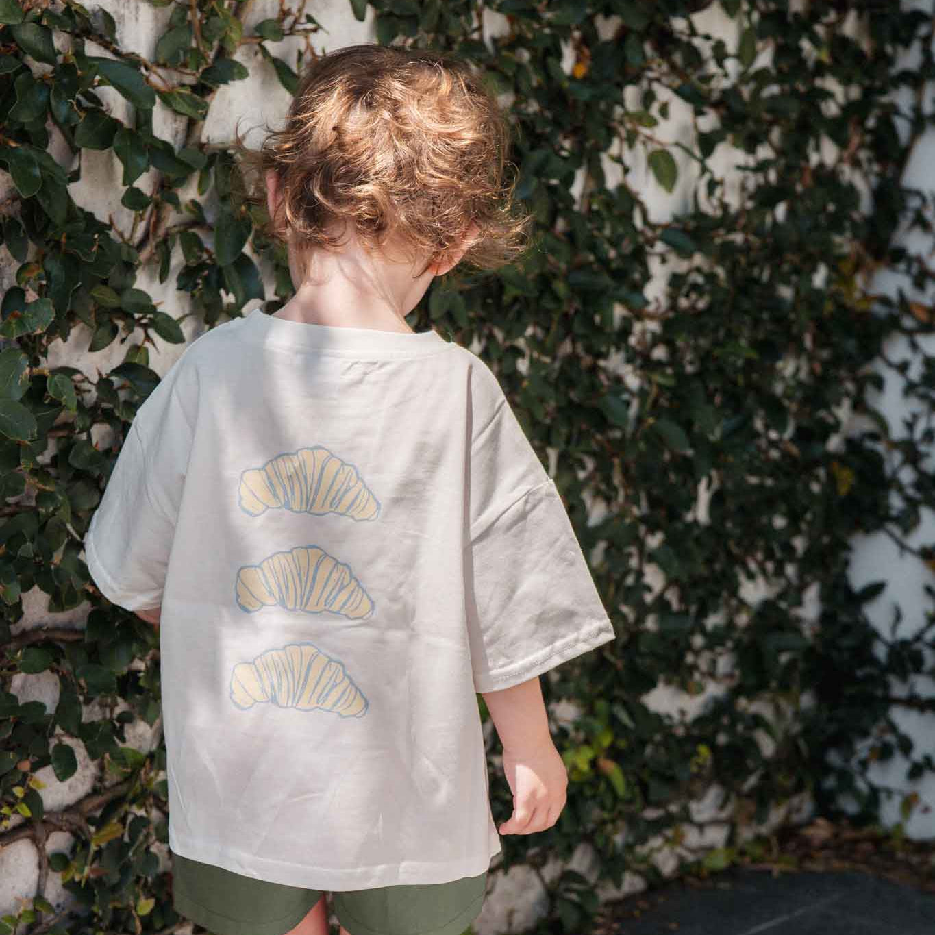 Child wearing a white t-shirt with croissant patterns and green shorts, standing against a leafy background.