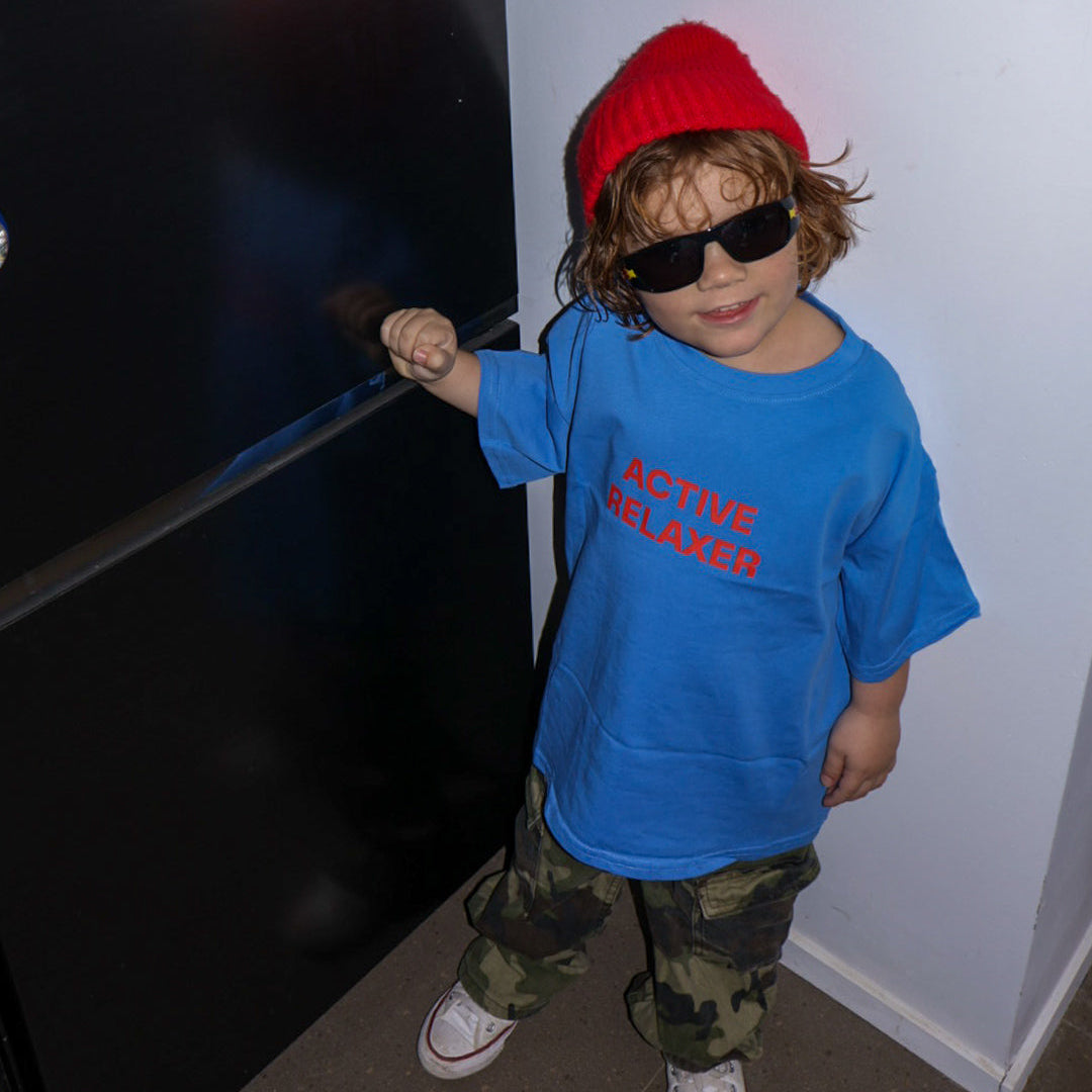 Child wearing a blue shirt with text, red beanie, and sunglasses standing in a room.