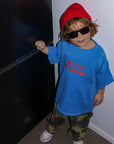Child wearing a blue shirt with text, red beanie, and sunglasses standing in a room.