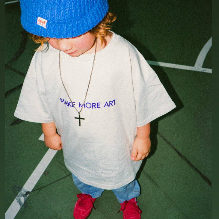 Child wearing a blue beanie and white t-shirt with 'MAKE MORE ART' text on a green floor.