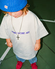 Child wearing a blue beanie and white t-shirt with 'MAKE MORE ART' text on a green floor.
