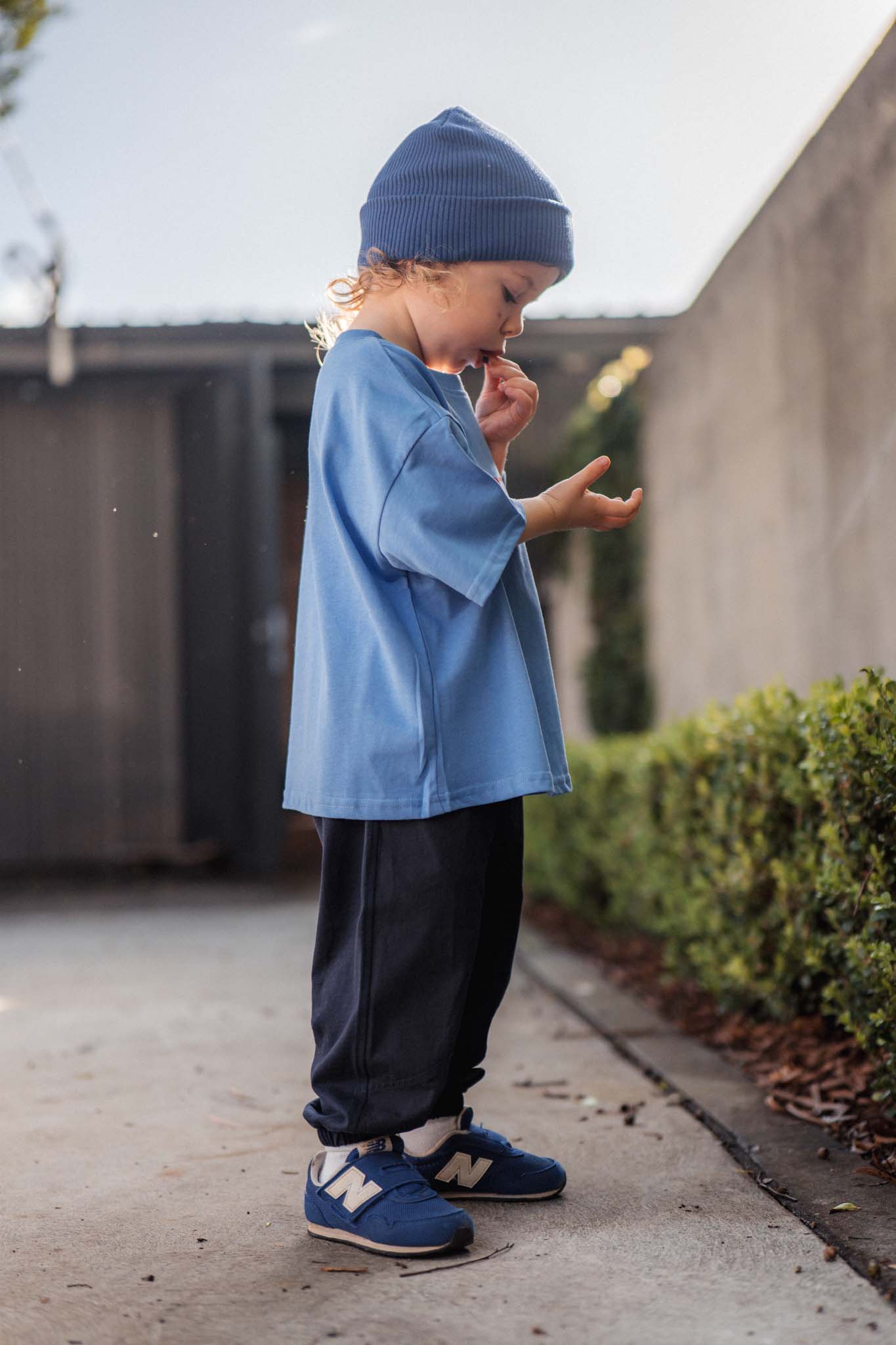Child wearing a blue outfit standing on a sidewalk with a building and greenery in the background