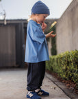 Child wearing a blue outfit standing on a sidewalk with a building and greenery in the background