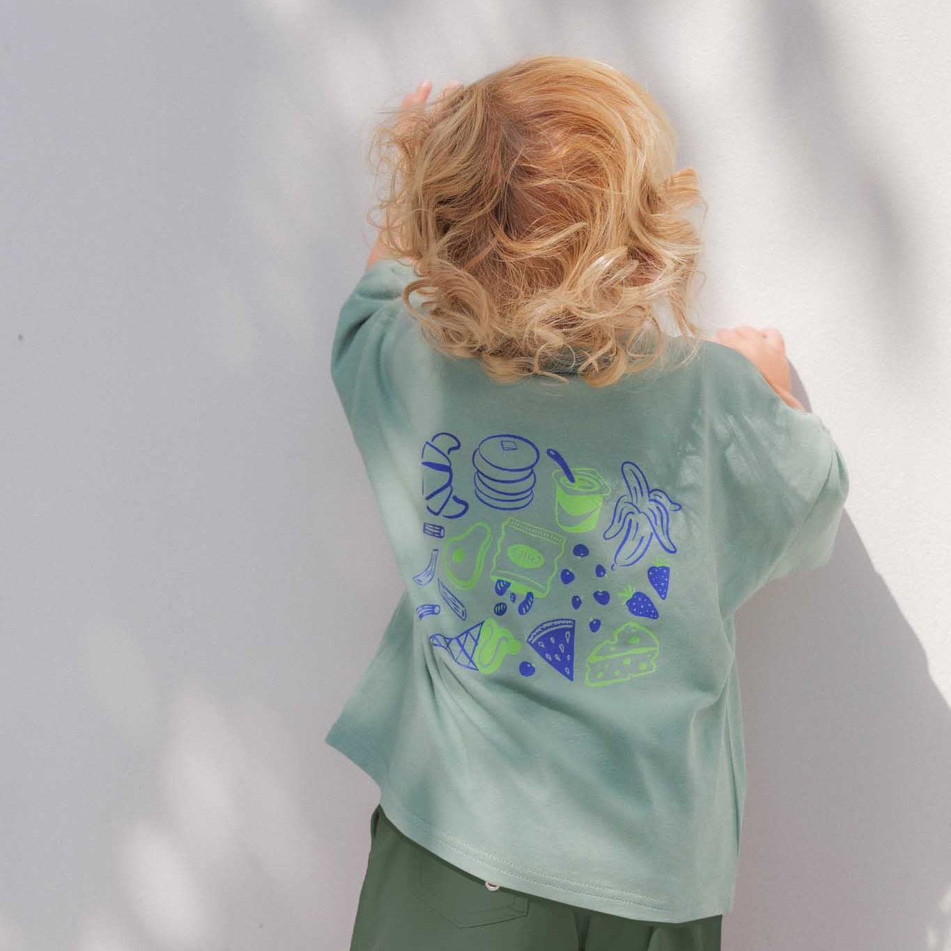 Child wearing a green snack lover tee with blue designs against a white wall