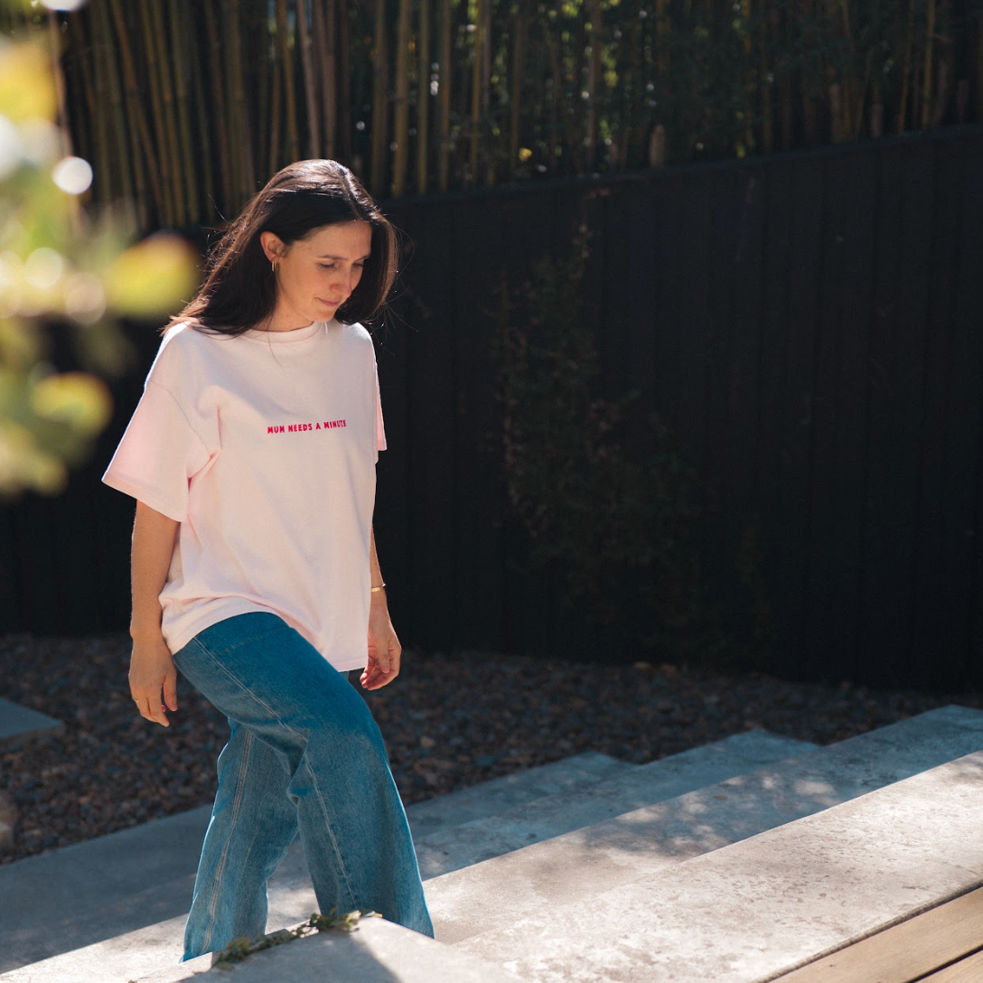 Woman wearing a pink t-shirt with red text and blue jeans standing on a stone ledge outdoors.