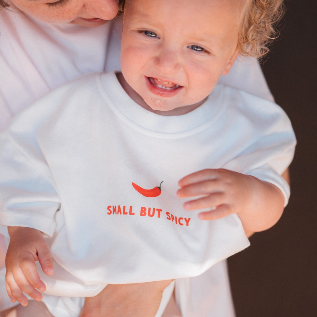 Child wearing a white shirt with red text and a red pepper graphic, held by an adult.