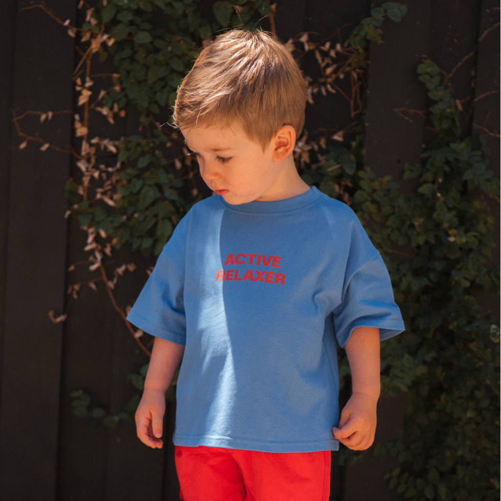 Young boy wearing a blue shirt with red text and red shorts standing on rocks with greenery in the background