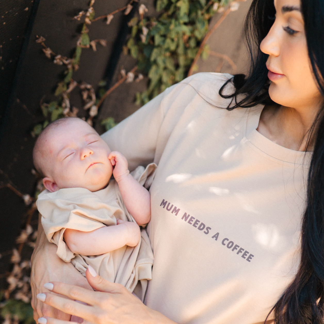 Woman holding a baby with 'Mum needs a coffee' shirt outdoors