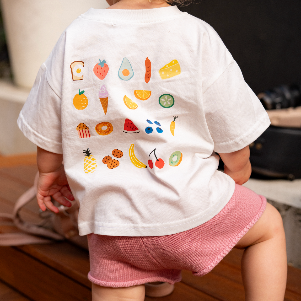 Child wearing a white t-shirt with colorful fruit illustrations and pink shorts.