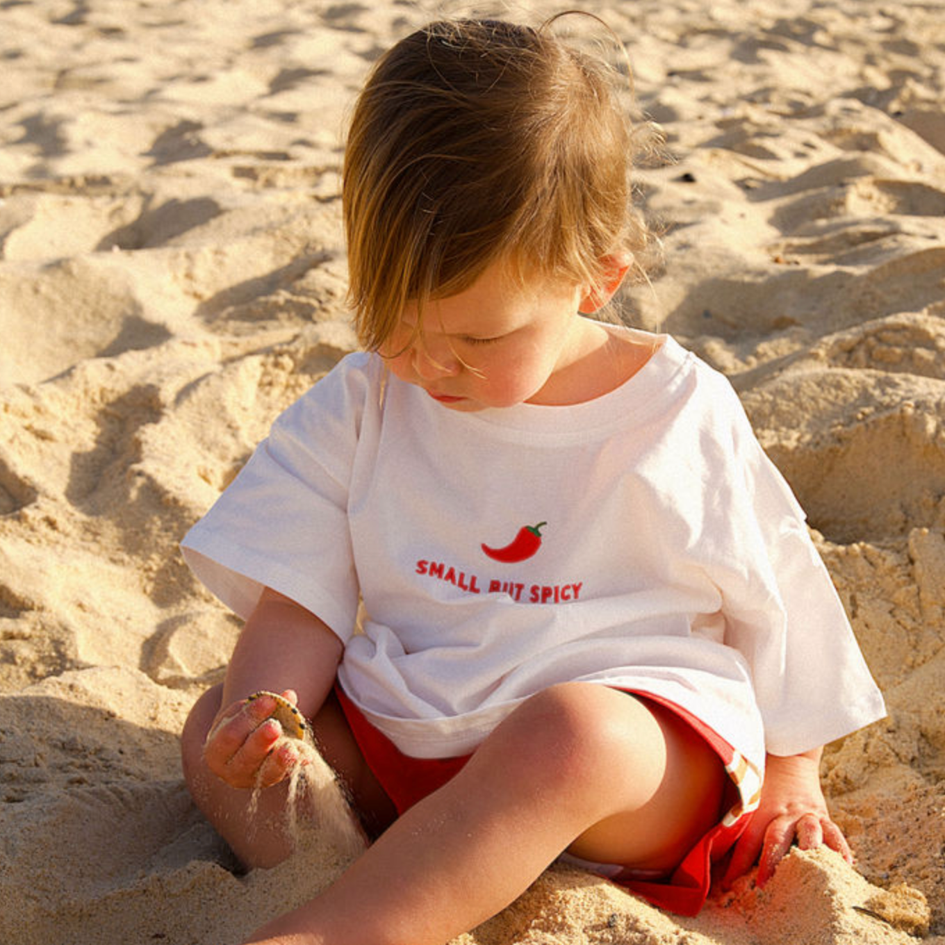 Child sitting on a sandy beach wearing a white t-shirt with text and red shorts.