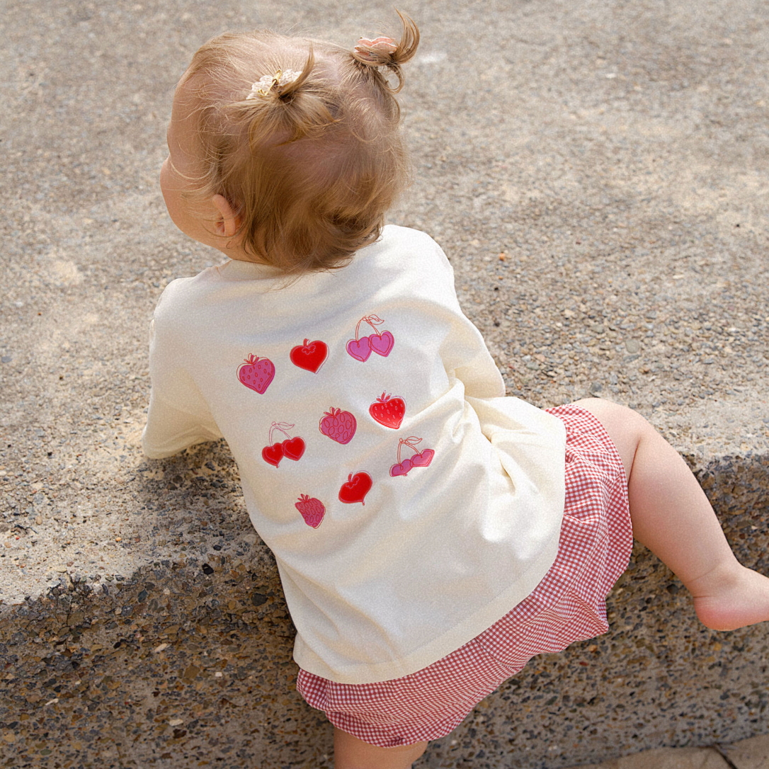 Child wearing a white shirt with red and pink heart patterns, sitting on a concrete surface.