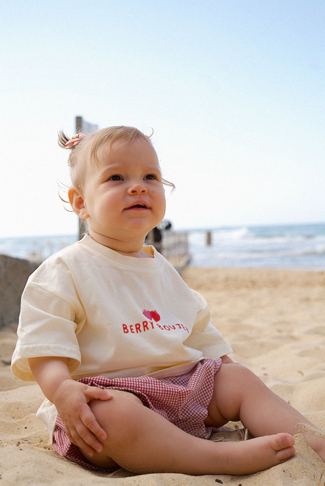 Child sitting on a sandy beach wearing a white shirt with red text and plaid shorts.