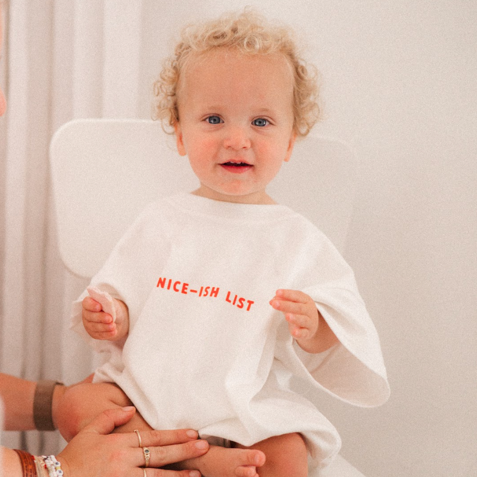 Child wearing a romoer with 'NICE-ISH LIST' text, sitting on a chair next to an adult.