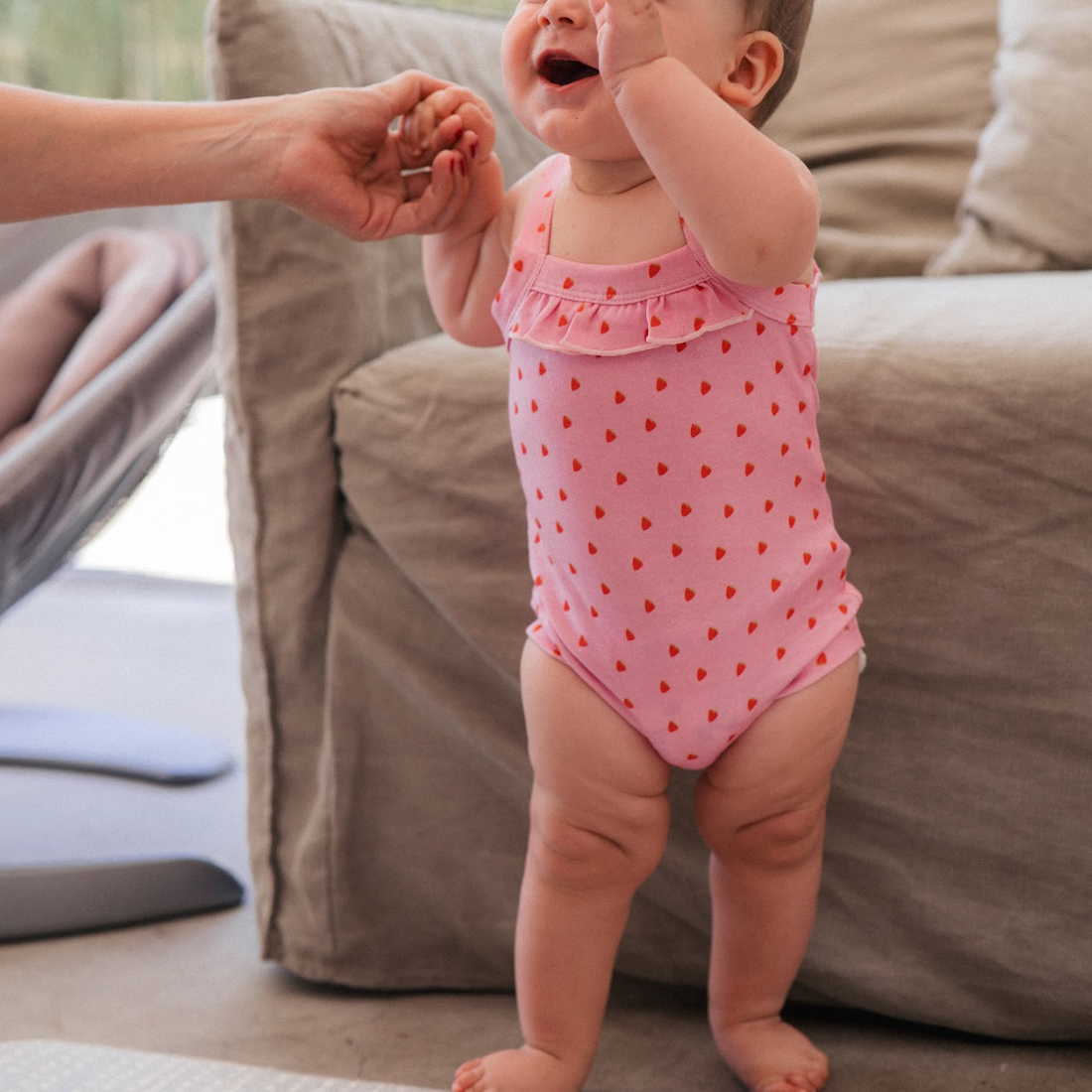 Baby in a pink swimsuit standing on a carpeted floor with a couch in the background.