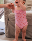 Baby in a pink swimsuit standing on a carpeted floor with a couch in the background.