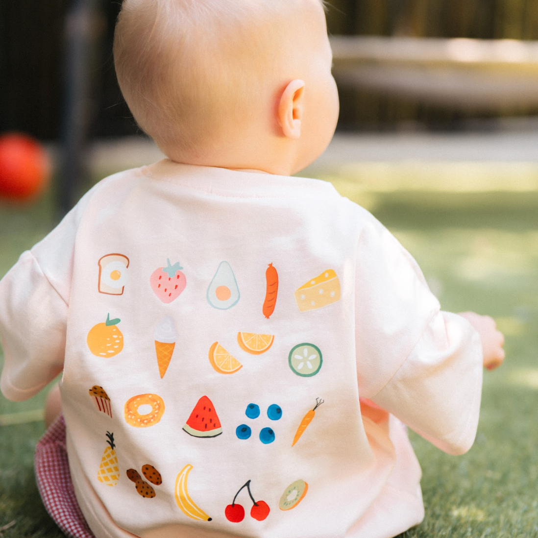 Baby wearing a white bib with colorful fruit and vegetable prints, standing on grass.