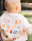 Baby wearing a white bib with colorful fruit and vegetable prints, standing on grass.