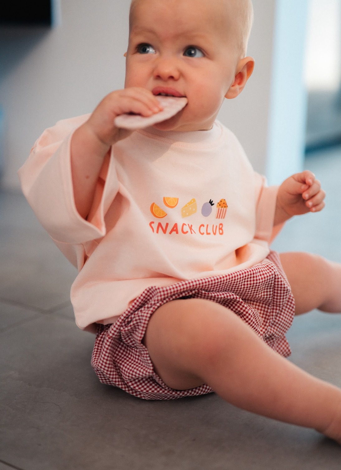 Baby wearing a pink 'Snack Club' shirt and plaid shorts, sitting on a floor.