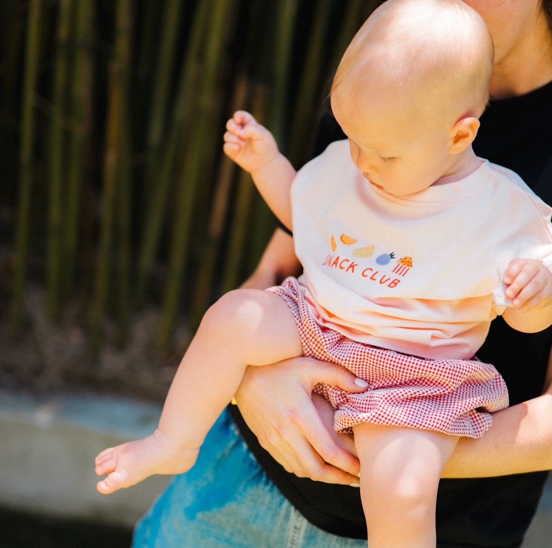 Baby wearing a 'Snack Club' shirt being held by an adult outdoors.