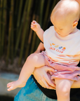 Baby wearing a 'Snack Club' shirt being held by an adult outdoors.
