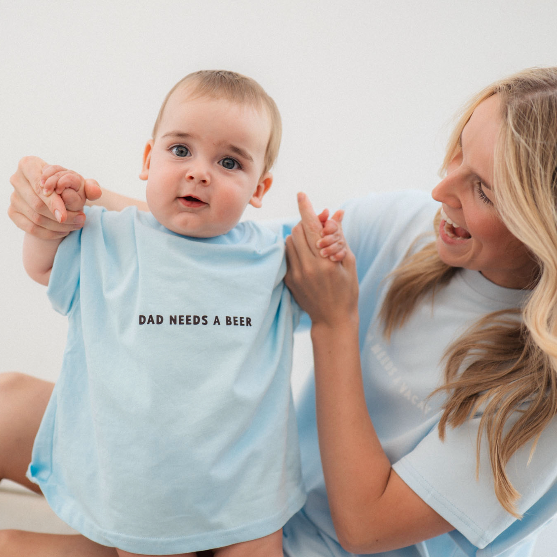 Woman holding a baby wearing a light blue onesie with 'DAD NEEDS A BEER' text.