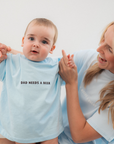 Woman holding a baby wearing a light blue onesie with 'DAD NEEDS A BEER' text.