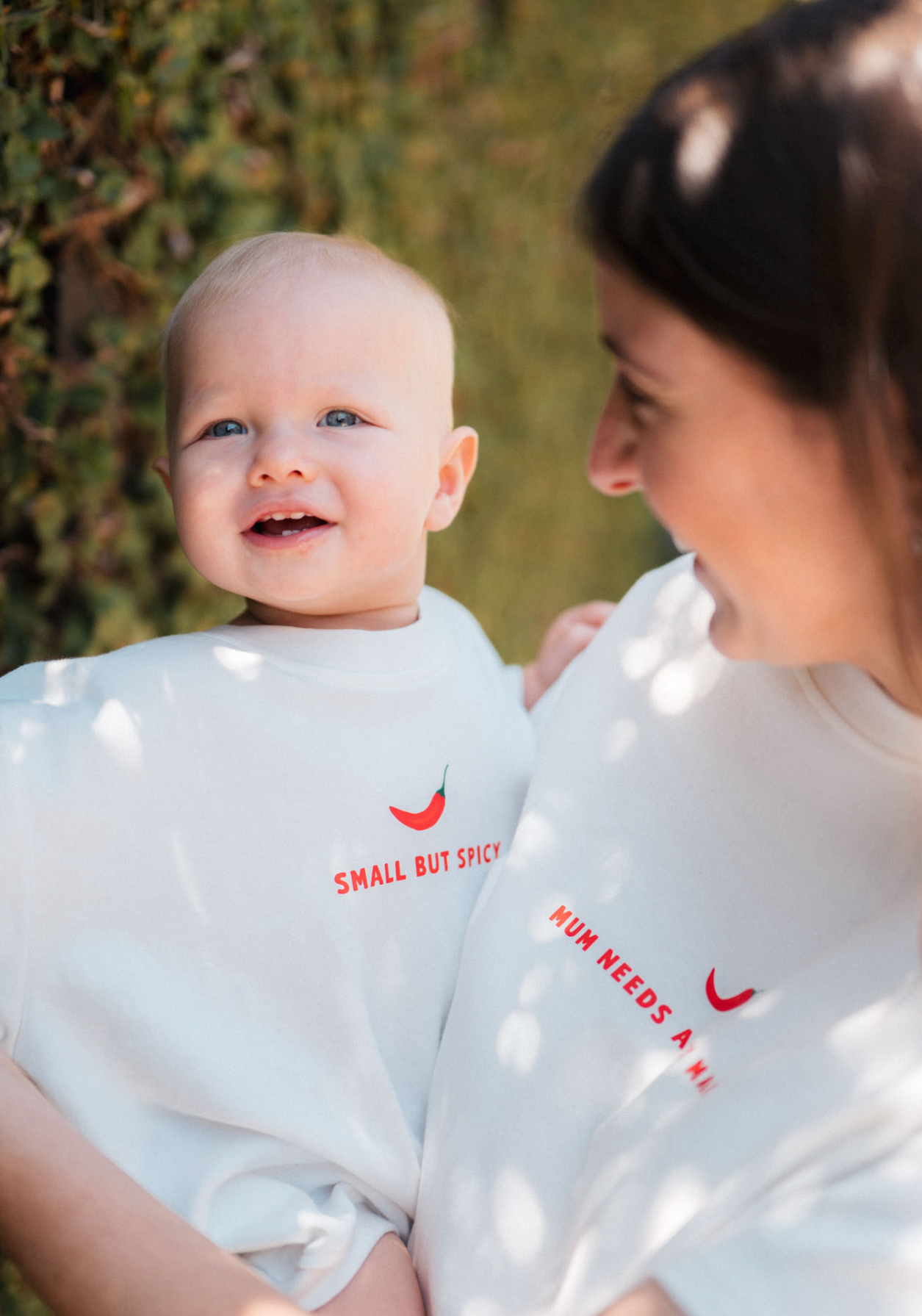 Woman and baby wearing matching white shirts with text in a natural setting