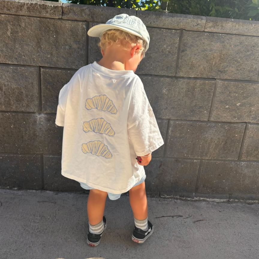 Child wearing a white t-shirt and cap standing against a stone wall.