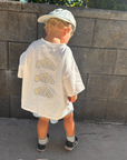 Child wearing a white t-shirt and cap standing against a stone wall.