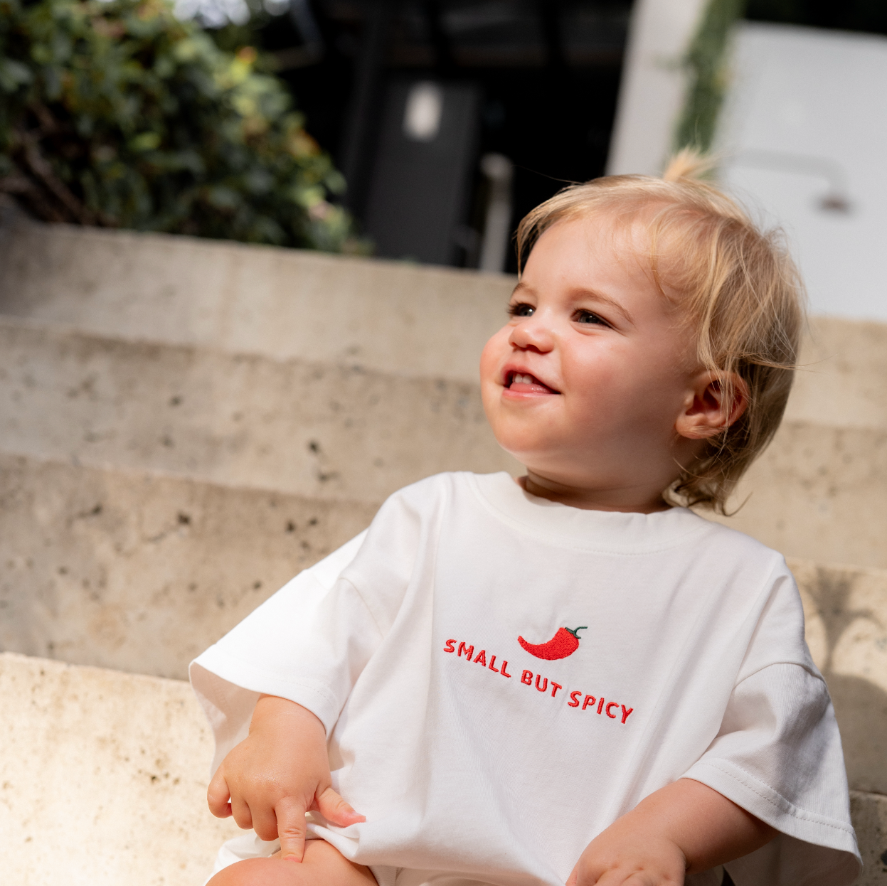 Child wearing a white t-shirt with 'Small But Spicy' text and red chili pepper graphic, sitting outdoors.