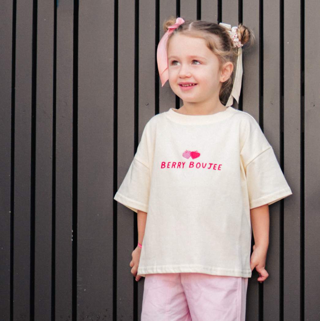 Child wearing a white t-shirt with berry boujee and pink pants standing against a dark vertical panel background