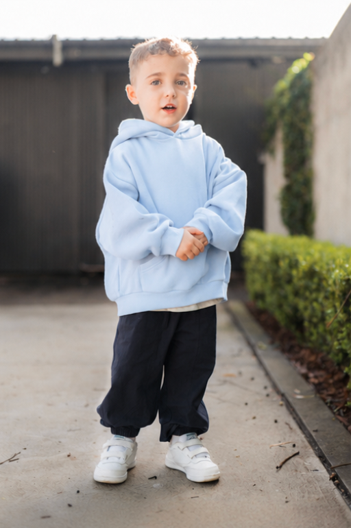 Child wearing a light blue hoodie and dark pants standing on a sidewalk.