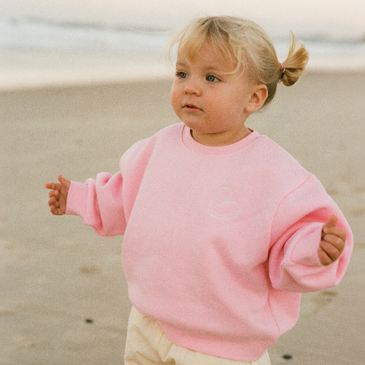 A toddler girl with blonde hair in a small ponytail stands on a sandy beach wearing a pink crewneck sweatshirt and cream-colored pants.