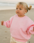 A toddler girl with blonde hair in a small ponytail stands on a sandy beach wearing a pink crewneck sweatshirt and cream-colored pants.