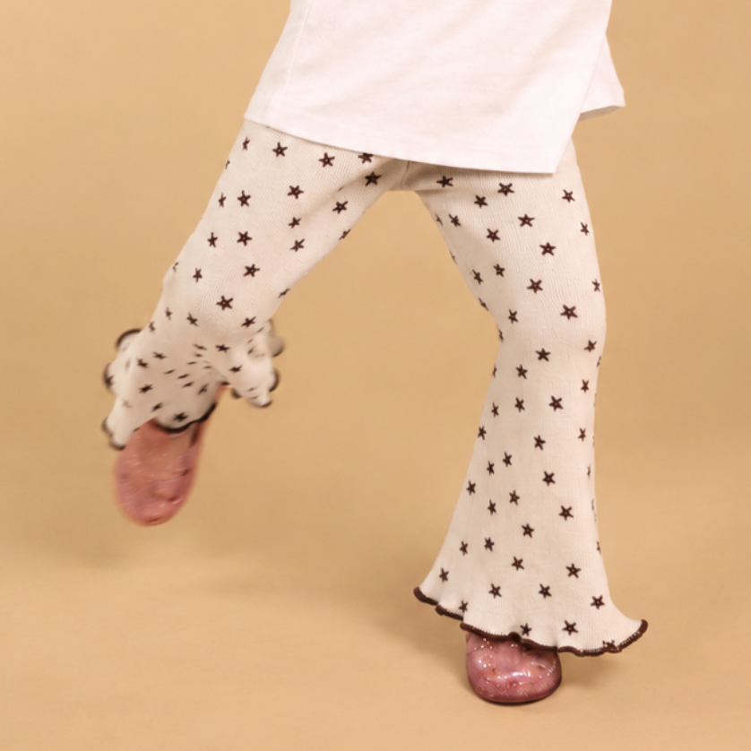 A child's lower body wearing cream flared pants with a black star pattern, a white t-shirt, and pink glittery shoes, captured mid-step against a solid beige background.