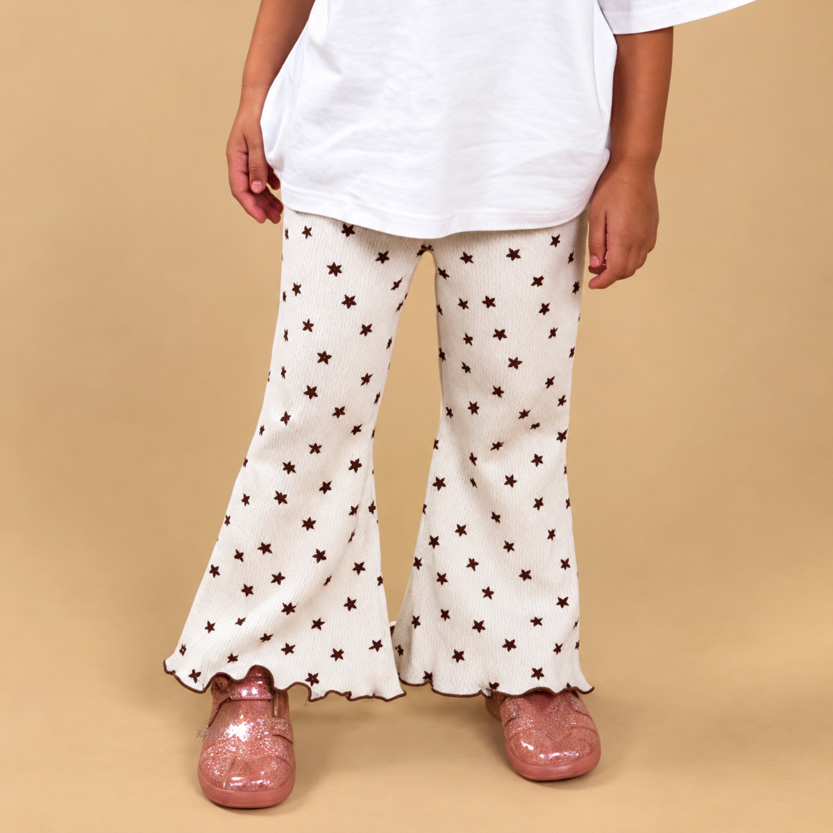 A child wearing cream flared pants with a brown star pattern, a white t-shirt, and pink clogs, standing against a solid beige background.