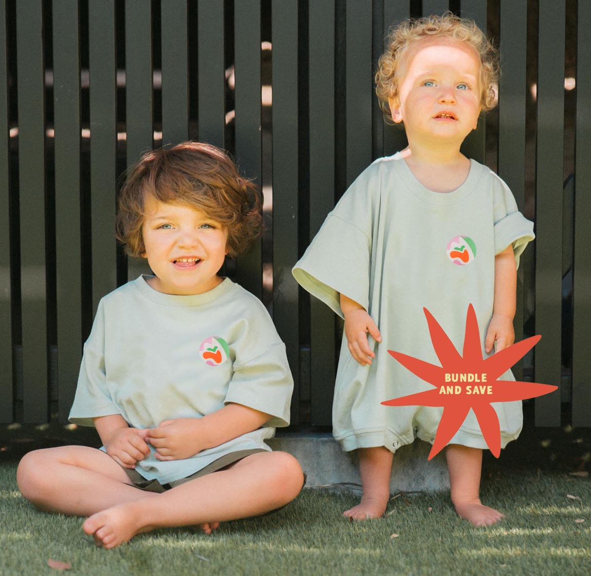 Two children wearing matching light green shirts with a logo, sitting on grass.