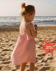 Young girl in a striped dress standing on a beach with ocean waves in the background.