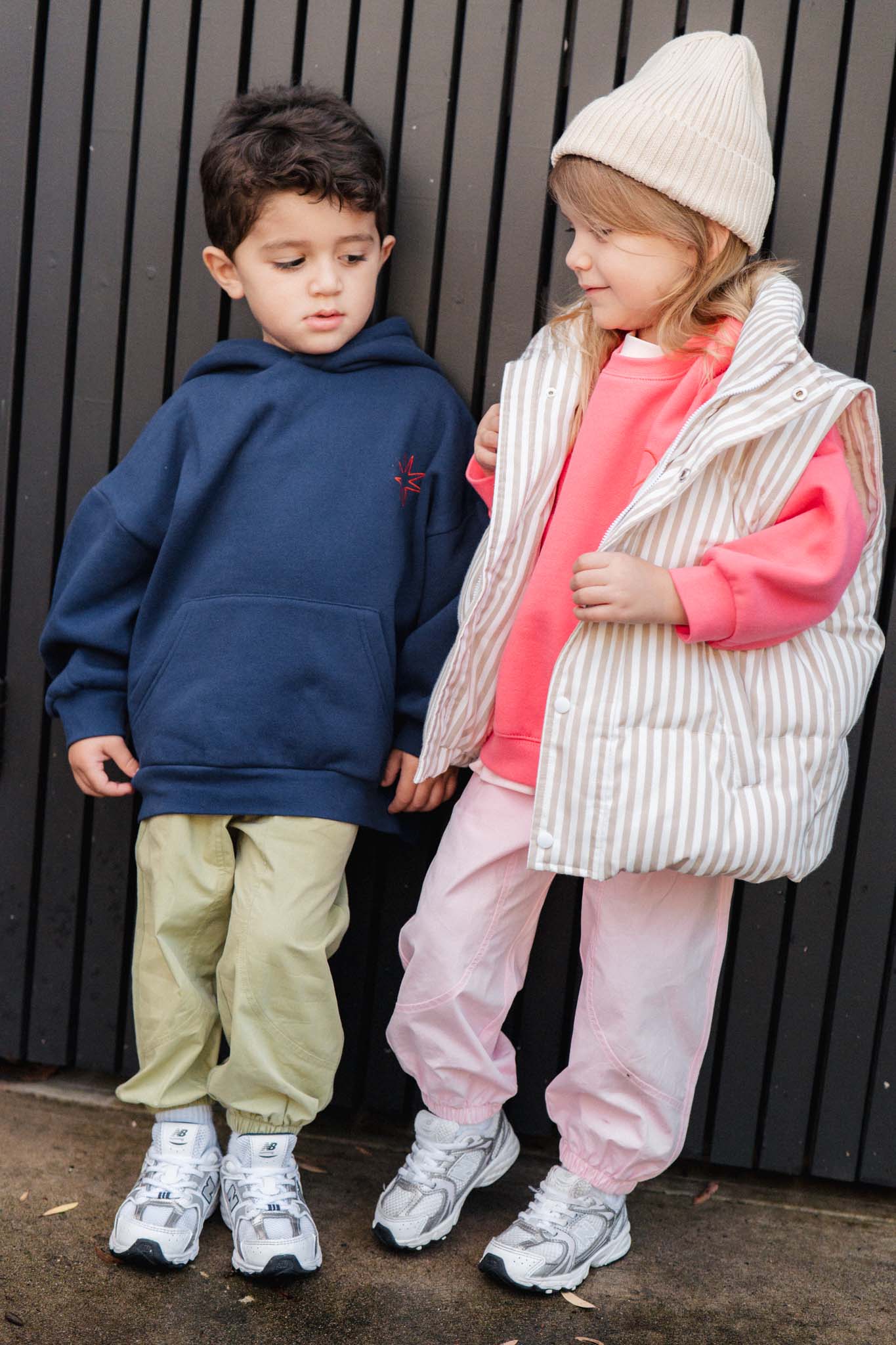 Two children standing against a dark background, wearing matching outfits with hoodies and pants.