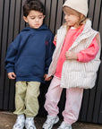 Two children standing against a dark background, wearing matching outfits with hoodies and pants.