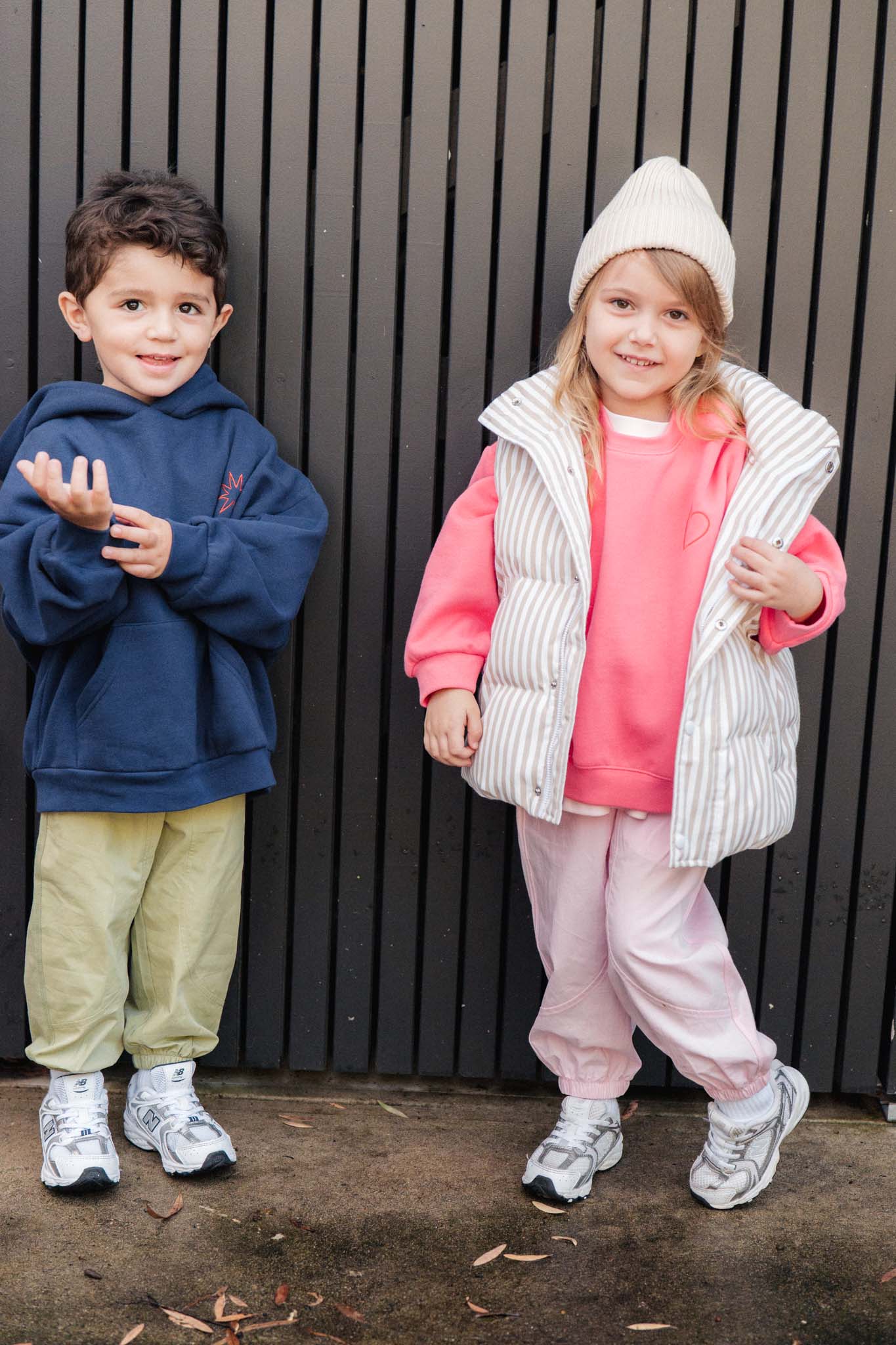 Two children standing against a black metal fence, one in a pink jumper with a striped puffer vest and pink pants.