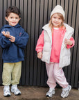 Two children standing against a black metal fence, one in a pink jumper with a striped puffer vest and pink pants.